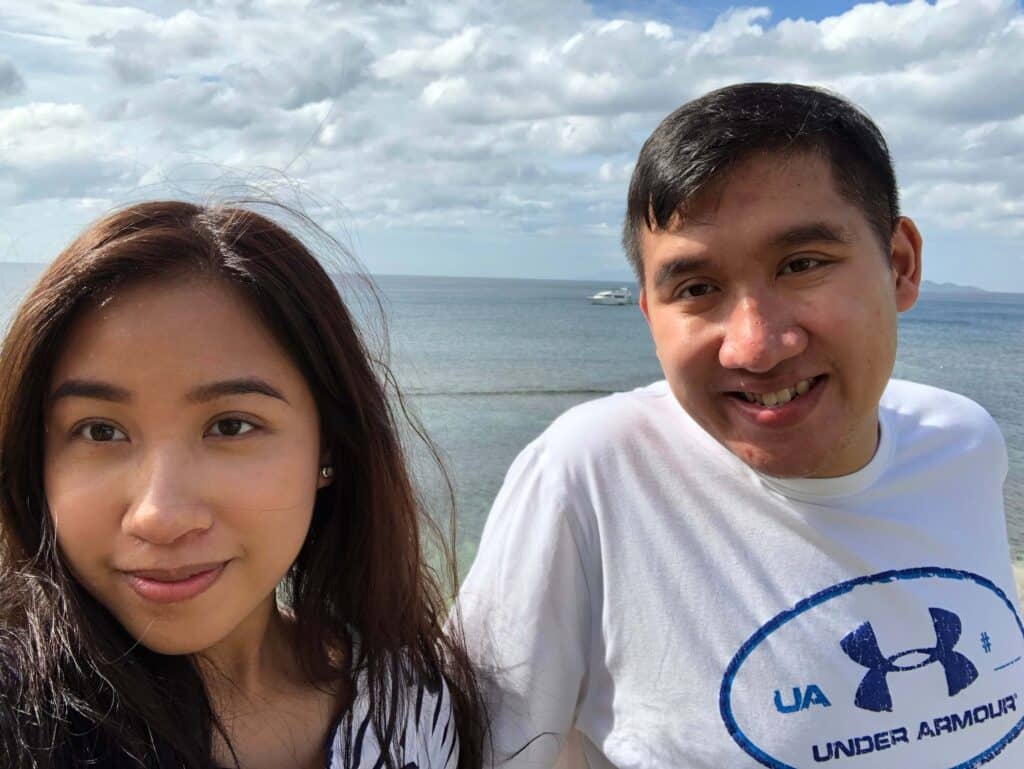 Photo of Melissa and her brother smiling in front of a cloudy sky and water.