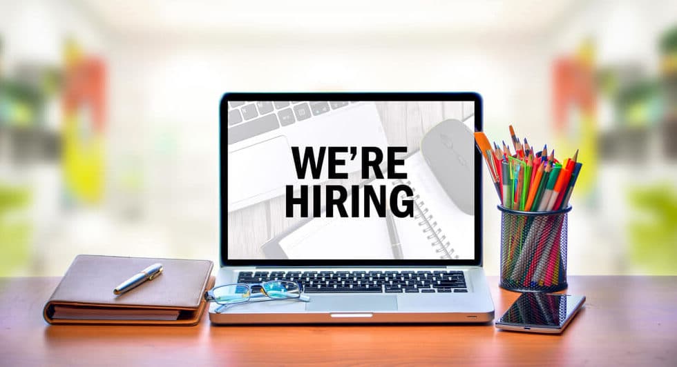 Image of laptop open on a desk that says "We're hiring." Next to the laptop is a notebook and a pencil cup.